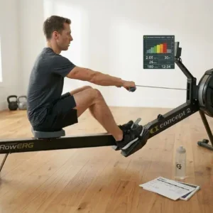 homme-utilisant-rameur-concept2-interieur-posture-prete-a Homme utilisant un rameur Concept2 en intérieur, posture prête à ramer, écran montrant des données, eau et doc à proximité. programme entraînement rameur débutant intégré naturellement.